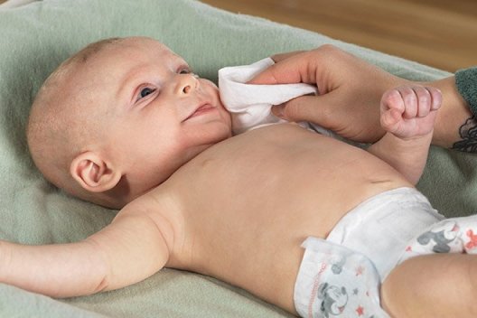 A parent uses a Huggies Natural Care Sensitive Wipe on their baby's face while they lay on a changing table
